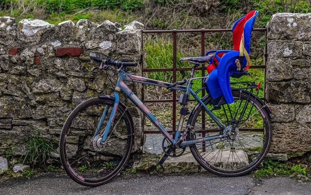 zaparkowany przy kamienno-metalowym ogrodzeniu rower trekkingowy z zamontowanym z tyłu czerwono-niebieskim fotelikiem dziecięcym
