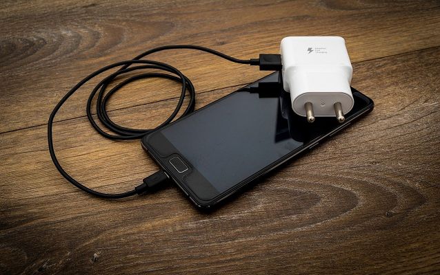 na drewnianej brązowej powierzchni leży czarny smarfon, do którego podpięty jest czarny kabel i biała ładowarka gniazdkowa