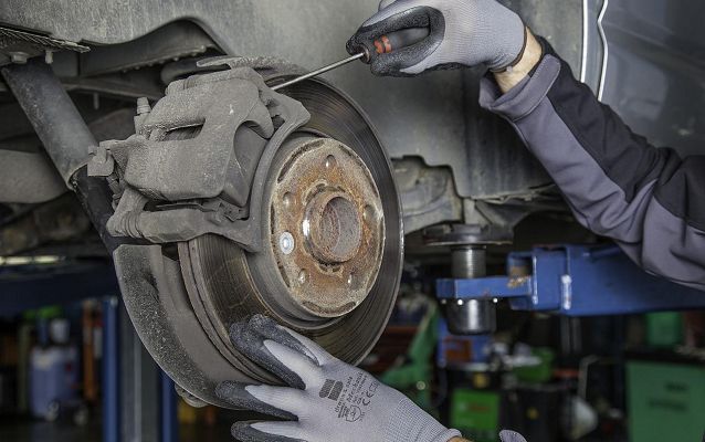 sprawdzenie tarcz hamulcowych na samochodzie znajdującym się wysoko na podnośniku. Na zdjęciu widać ponadto ręce mechanika sprawdzającego poszczególne elementy hamulca