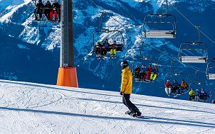 Stok narciarski, z którego zjeżdża na desce snowboardowej osoba ubrana w żółtoczarny strój narciarski. W dalszej części zdjęcia wyciąg krzesełkowy, zajęty przez narciarzy, a w tle ośnieżone stoku gór. Obraz Ri Butov z Pixabay
