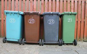 Cztery pojemniki na śmieci na kółkach ułatwiających ich opróżnienie, w różnych kolorach - w kolejności od lewej: niebieski, brązowy, grafitowy i zielony. Stoją na betonowym podłożu, na tle jasnobrązowwego płotu. Image by Davie Bicker from Pixabay